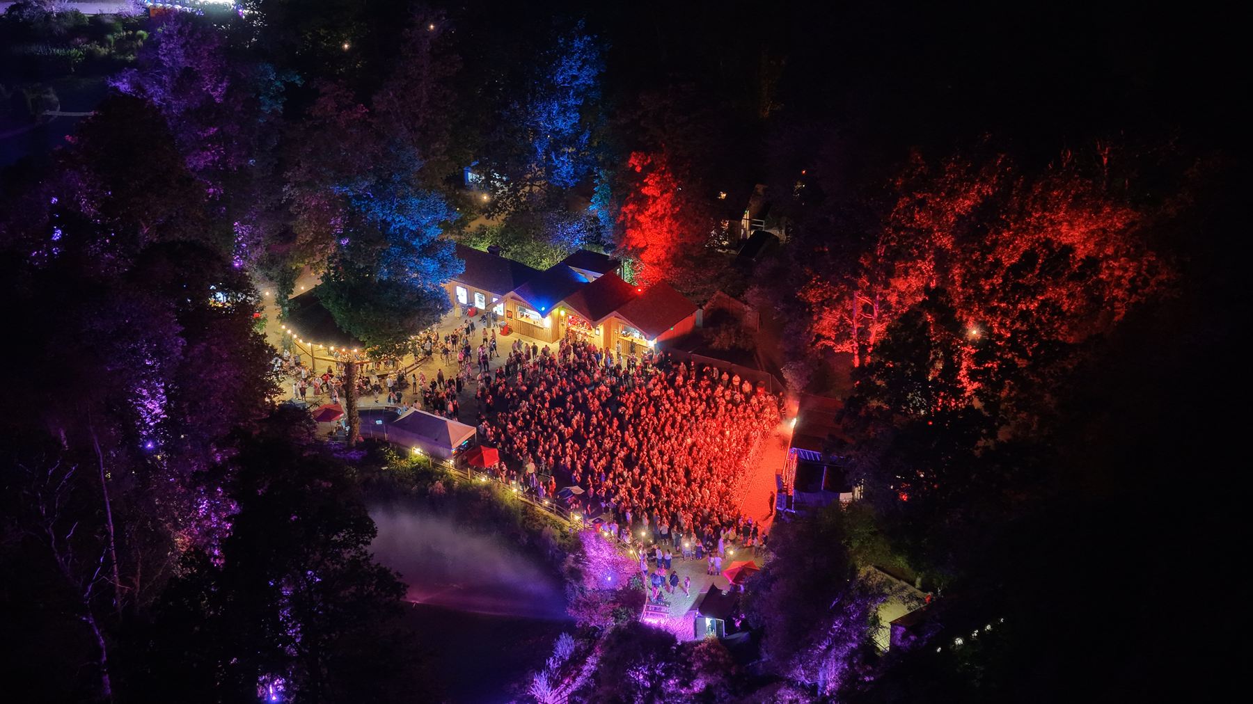 Night aerial photograph of a woodland festival arena, the crowd packed between lit buildings with the trees washed red and blue