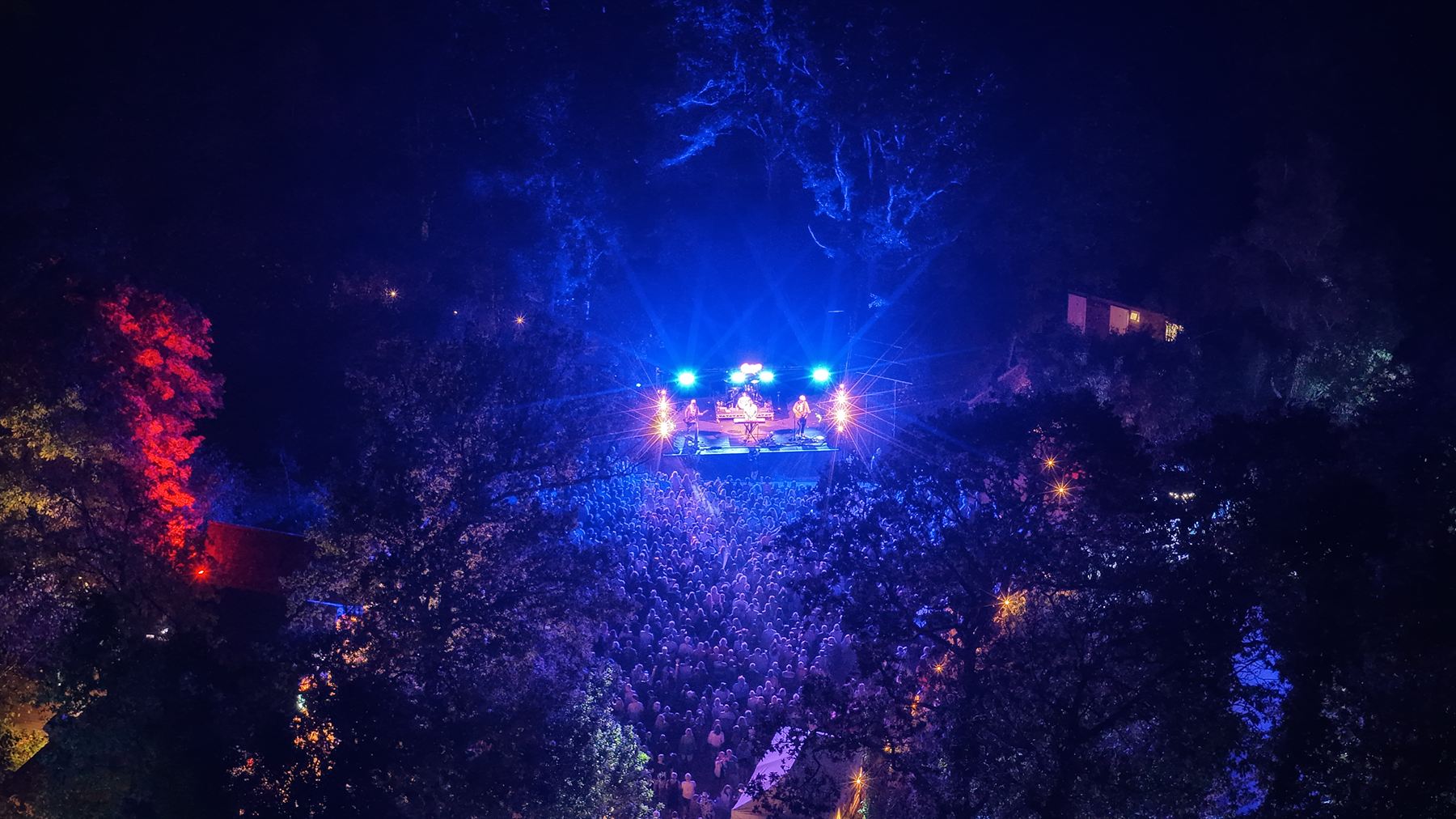 Aerial view through tree canopy of the Strings Festival crowd lit blue in front of the stage