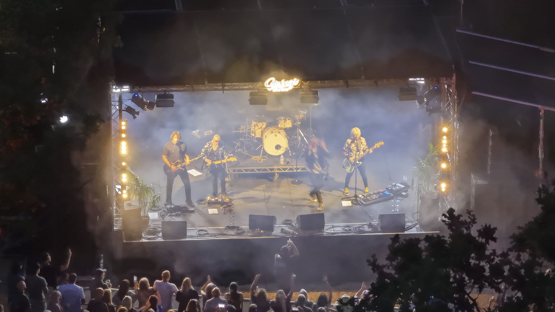 Close aerial view of the Strings Festival stage, a four-piece band performing under warm lighting and haze with the crowd in the foreground